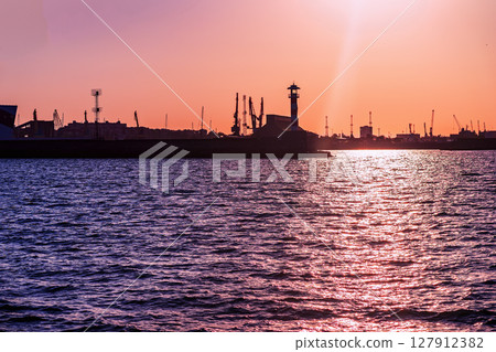 Vibrant orange sunset illuminates port skyline with cranes and striped lighthouse reflected in sea. Concept of lighthouse, industrial coast, colorful maritime industrial background Vibrant orange sunset illuminates port skyline with cranes and striped lighthouse reflected in sea. Concept of lighthouse, industrial coast, colorful maritime industrial background 127912382