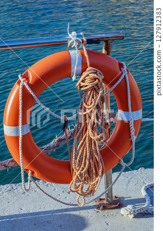 Bright orange lifebuoy with attached rope hangs from metal rail above clear coastal water for emergency use. Concept of lifebuoy, safety gear, marine protection Bright orange lifebuoy with attached rope hangs from metal rail above clear coastal water for emergency use. Concept of lifebuoy, safety gear, marine protection 127912383