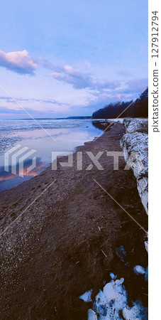 Spring landscape. Panoramic view of beautiful dawn on bay. Cumulus clouds over water in bright light. Ice, snow and rocks on coastline. Rising rays of sun are reflected in sea. Vertical photo. 127912794
