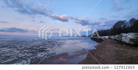 Spring landscape. Panoramic view of beautiful dawn on bay. Cumulus clouds over water in bright light. Ice, snow and rocks on coastline. Rising rays of sun are reflected in sea. High quality photo 127912803