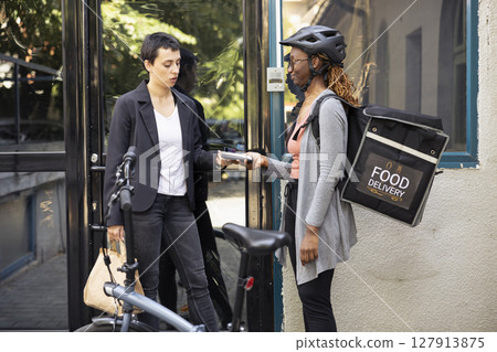 Woman completes a fast food purchase with contactless payment at the doorstep, courier holds pos terminal steady as the electronic transaction is processed. Electronic wireless purchase. 127913875