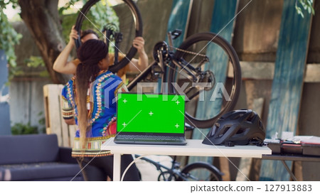 Laptop showing greenscreen while healthy and active interracial pair fixes bicycle components. Minicomputer with isolated copyspace screen on table while couple does bike maintenance at home yard. Laptop showing greenscreen while healthy and active interracial pair fixes bicycle components. Minicomputer with isolated copyspace screen on table while couple does bike maintenance at home yard. 127913883