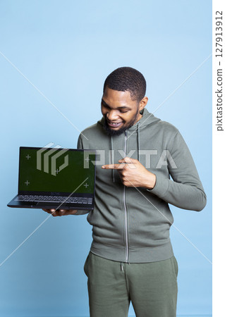 Confident happy person pointing at green screen mockup in studio, using personal laptop for an advertisement on camera. African american young guy gesturing against blue background. 127913912