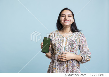 Indian woman showing isolated copy space display in front of the camera, creating an advertisement for a commercial against blue background. Young joyful adult holding device in studio. Indian woman showing isolated copy space display in front of the camera, creating an advertisement for a commercial against blue background. Young joyful adult holding device in studio. 127913919