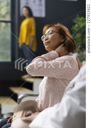 Physician evaluating neck injury and muscle ache during diagnosis checkup in a modern hospital lobby. Asian woman describing pain discomfort while a healthcare worker suggests therapy options. 127913921