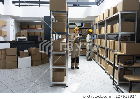 Woman employee conducting manual labor surrounded by packages and crates, staff managing shipping, delivery and inventory control tasks. Warehouse interior supply and demand operations. Woman employee conducting manual labor surrounded by packages and crates, staff managing shipping, delivery and inventory control tasks. Warehouse interior supply and demand operations. 127913924