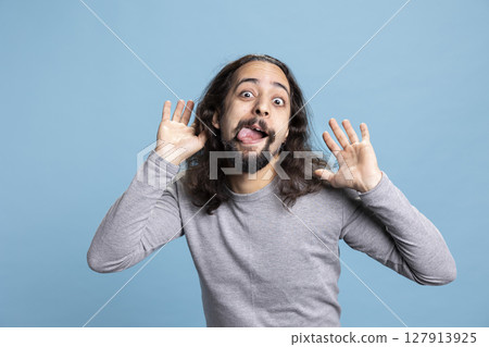 Playful young man with a long hair and beard making a goofy face in the studio. Carefree guy radiates humor and joy, capturing a lighthearted moment full of laughter and casual personality. Playful young man with a long hair and beard making a goofy face in the studio. Carefree guy radiates humor and joy, capturing a lighthearted moment full of laughter and casual personality. 127913925