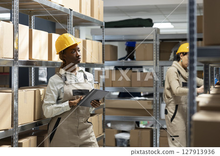 Black woman oversees delivery processes and controlling inventory, checking boxes in a warehouse setting. Handling industrial workspace dedicated to product management and logistics solutions. Black woman oversees delivery processes and controlling inventory, checking boxes in a warehouse setting. Handling industrial workspace dedicated to product management and logistics solutions. 127913936