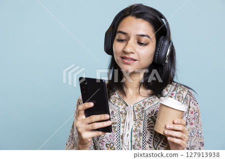 Pleased excited woman playing music on phone and drinking coffee, having fun dancing on her favorite songs against blue background. Delighted indian girl wearing headphones and doing funky moves. 127913938