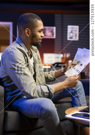 Young freelancer accountant doing math calculations using financial graphs on papers, working from his cozy home. Self employed person comparing business economic figures on files. 127913939