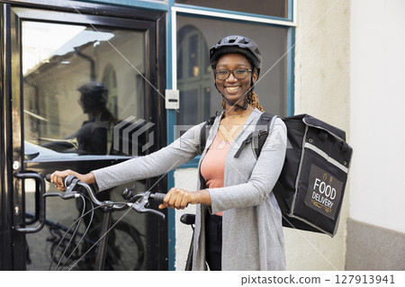 Portrait of smiling black woman working as a takeaway courier on the bike, riding with a helmet and carrying her backpack. Takeout employee delivering fast food or other meal orders. 127913941