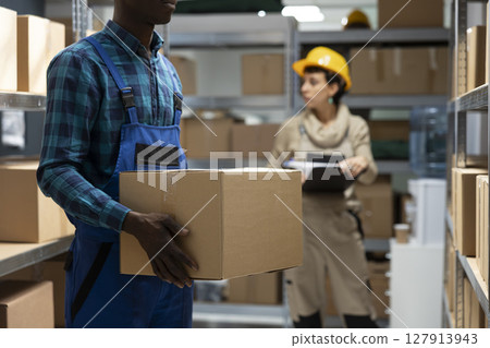 Industrial depot environment focused on import export efficiency and storage. Black employee handles manual work tasks such as inventory tracking, goods handling and shipment preparation. Industrial depot environment focused on import export efficiency and storage. Black employee handles manual work tasks such as inventory tracking, goods handling and shipment preparation. 127913943
