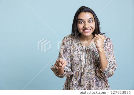 Satisfied joyous person cheering for something and clenching his fists, being ecstatic and overjoyed about good news or achievements. Joyful optimistic girl feeling surprised against studio background 127913955