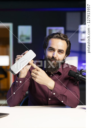 Joyful influencer doing technology review of wireless portable speaker for internet platforms channel. Influencer pointing to music playing gear to his subscribers, recommending it Joyful influencer doing technology review of wireless portable speaker for internet platforms channel. Influencer pointing to music playing gear to his subscribers, recommending it 127913957