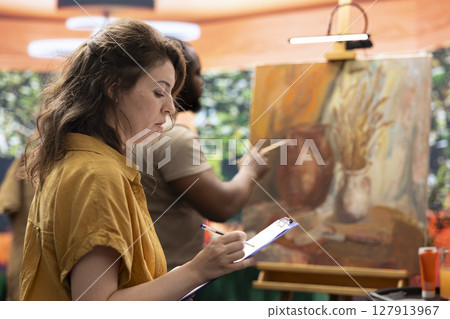 Skilled master painter keeps account of her apprentice techniques, following his creative process and teaching him new ideas. Beginner artist listening to advice from experienced woman. 127913967