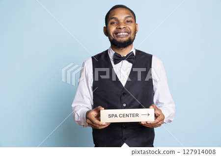 Hotel porter in suit and tie helping with directions to the spa center using a pointer, holding the wall sign to guide guests to resort amenities. Providing great concierge services. 127914008