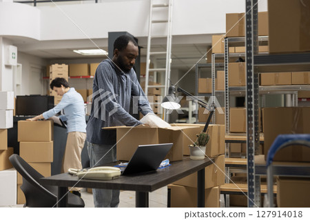 Black male executive manager uses laptop for order logistics in the storage room, working in a local distribution hub. Supply room filled with boxes and inventory goods, e commerce. 127914018