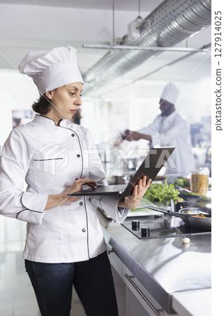 Dedicated woman chef using laptop for research and tips in cooking gourmet meals, restaurant kitchen balancing technology and tradition. Expert chopping vegetables and raw materials. Dedicated woman chef using laptop for research and tips in cooking gourmet meals, restaurant kitchen balancing technology and tradition. Expert chopping vegetables and raw materials. 127914025