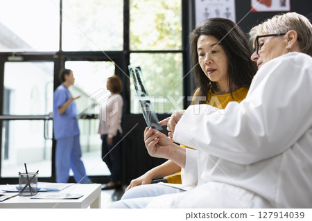 General practitioner analyzing X ray test results with asian woman in a lobby, providing guidance and a treatment plan. Hospital specialist working in public health industry, medical care. 127914039