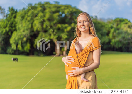 Mother holding her little baby in a yellow sling in the park. Warm and loving family moment. Babywearing, parenting, and mother-child bonding concept Mother holding her little baby in a yellow sling in the park. Warm and loving family moment. Babywearing, parenting, and mother-child bonding concept 127914185