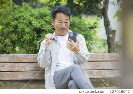 一位老人坐在公園的長椅上看著他的智慧型手機 127916042