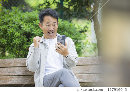 一位老人坐在公園的長椅上看著他的智慧型手機 127916043