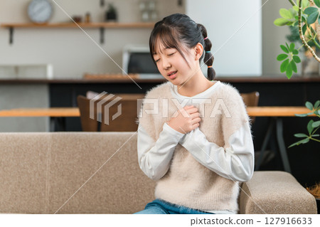 A girl in elementary and middle school sitting on a sofa at home holding her chest due to chest pain A girl in elementary and middle school sitting on a sofa at home holding her chest due to chest pain 127916633