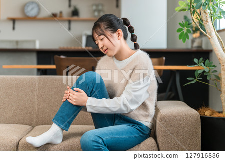 A girl in elementary and middle school sitting on a sofa at home holding her knees and shins due to knee pain 127916886