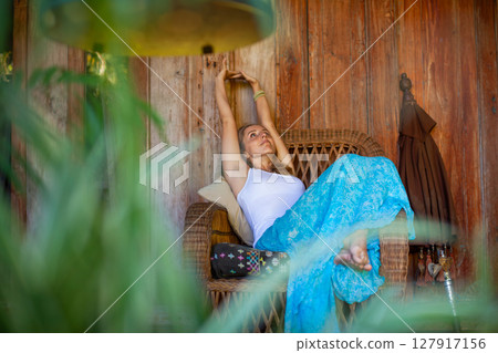 Young woman sit on open-air veranda of wooden bungalow with tropical garden view. Girl relaxing in luxury villa on family summer vacation. Countryside lifestyle. Young woman sit on open-air veranda of wooden bungalow with tropical garden view. Girl relaxing in luxury villa on family summer vacation. Countryside lifestyle. 127917156