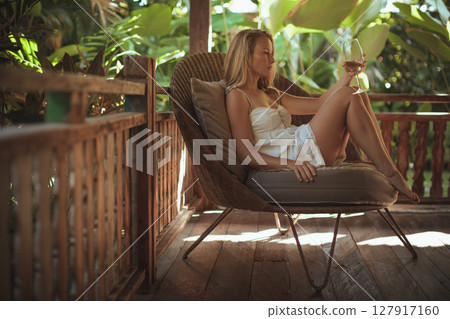 Young woman sit on open-air veranda of wooden bungalow with tropical garden view. Girl relaxing in luxury villa on family summer vacation. Tropical lifestyle. Young woman sit on open-air veranda of wooden bungalow with tropical garden view. Girl relaxing in luxury villa on family summer vacation. Tropical lifestyle. 127917160