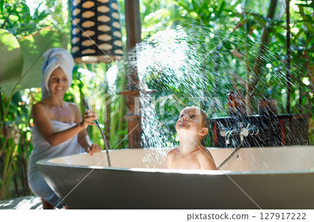Happy baby son with mother have fun in bath. Playful woman spraying child from shower in outside bathroom on open veranda with beautiful tropical garden view in luxury villa on summer family vacation. 127917222