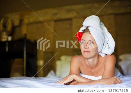 Relaxing woman with towel and red flower on head lies on bed thinking. Spa morning, beauty care, peaceful self-care time, skincare, wellness retreat, calm lifestyle, hotel relaxation. 127917226