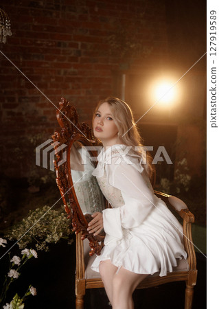 Blonde woman in white dress sitting by vintage mirror in moody room with brick wall background Blonde woman in white dress sitting by vintage mirror in moody room with brick wall background 127919589