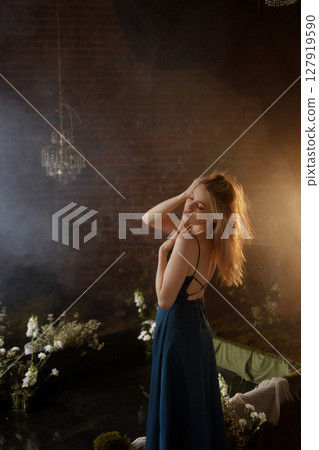 Woman in blue dress posing under vintage chandelier in dark room with brick wall and flowers 127919590