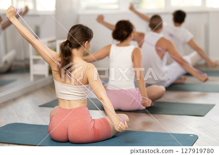 Back view of young girl with group of young adults maintaining physical health practicing pilates with resistance band sitting on gymnastics mat at fitness center 127919780