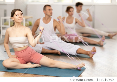 Active young girl wearing sport clothes using resistance band during group workout sitting on mat in gym 127919792