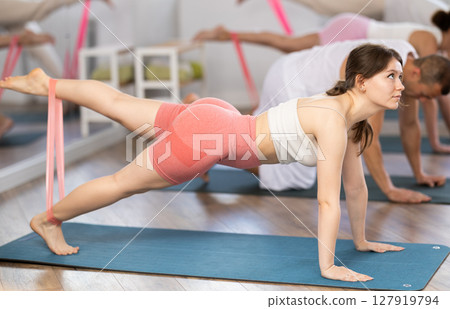 Athletic sporty young girl on hands and knees doing stretch exercises for legs with resistance bands taking part in intense group training in gym 127919794