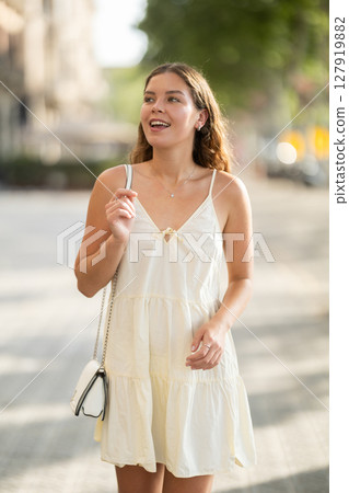 Cheerful girl in white dress walking along the street among architecture with bag 127919882