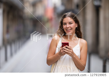Happy young woman with mobile phone in her hands walking around city and taking pictures of sights Happy young woman with mobile phone in her hands walking around city and taking pictures of sights 127919905