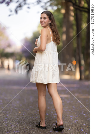 Cheerful girl in white dress walking along the street among architecture with bag 127919950