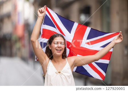 Cheerful girl holding national flag of UK flag, standing outdoors Cheerful girl holding national flag of UK flag, standing outdoors 127920001
