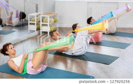 Young men and women practicing leg lift with band on knees lying on the side 127920256