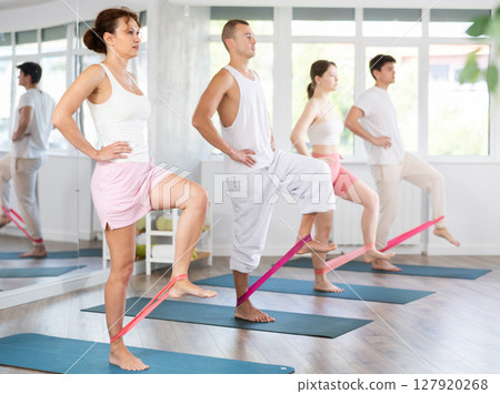 Young men and women practicing hip workout with band standing on mat 127920268