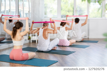 Young men and women doing shoulder workout with resistance band sitting cross-legged 127920276