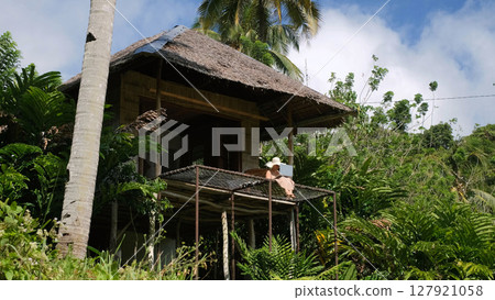 Young woman in a straw hat focuses on her laptop, seated at a bamboo hut terrace surrounded by lush jungle greenery, freelance and remote work in scenic natural setting  127921058