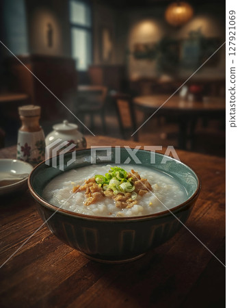 Delicious rice porridge with pork and green onions served in a ceramic bowl on a wooden table, with a blurred restaurant background. 127921069