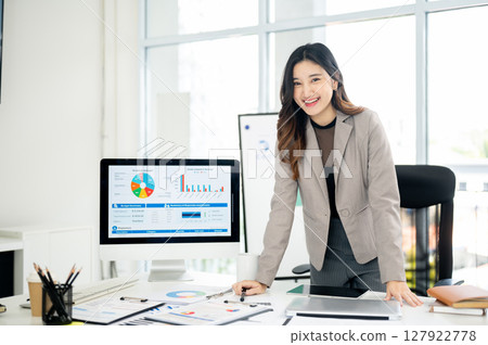 Businesswoman or female office worker is leaning over table next to monitor showing data and charts. Businesswoman or female office worker is leaning over table next to monitor showing data and charts. 127922778