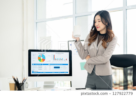 Businesswoman or female office worker holding mug standing next to monitor showing charts and data. 127922779