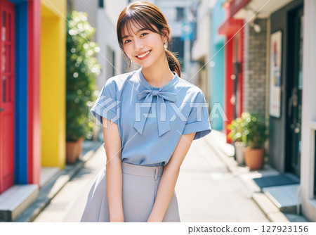 A female model wearing a light blue blouse and gray skirt, smiling and standing in a city lined with colorful buildings on a summer day 127923156
