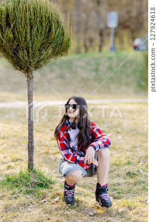 Young skater girl crouching in a park wearing headphones and sunglasses 127924618